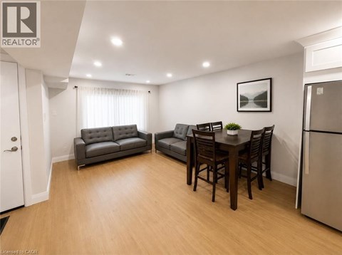 A well-lit living room with a grey sofa, a black leather armchair, a wooden dining table with chairs, and a refrigerator.
