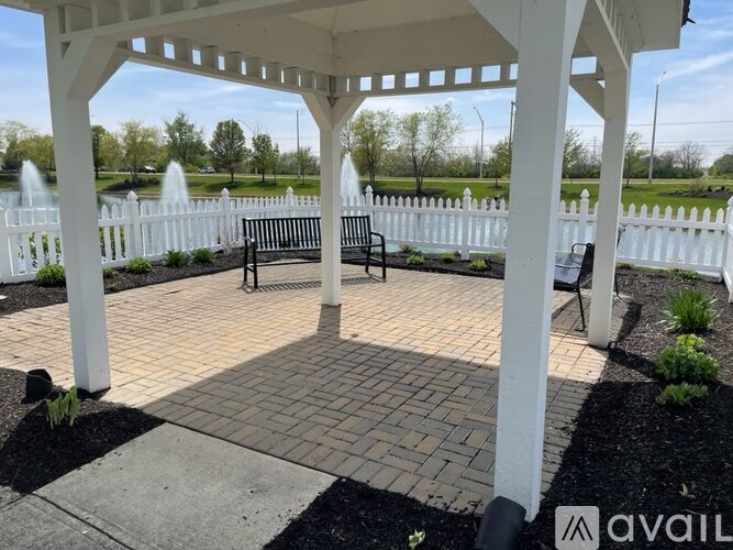 A white gazebo with a bench in front of a white picket fence and a water feature in the background.