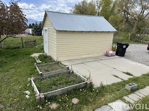 A small beige building with a metal roof is surrounded by a garden bed and a trash can.