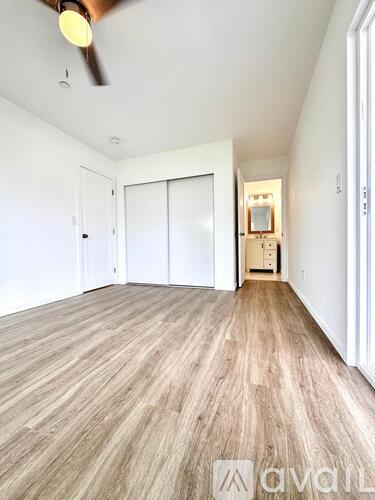 A room with a ceiling fan and light, wooden flooring, and white walls with a doorway leading to another room.