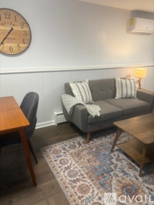 A grey couch with pillows sits in a living room with a wooden coffee table and a clock on the wall.