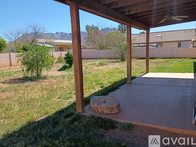 A patio with a wooden roof and concrete floor is surrounded by a grassy area and a fence.