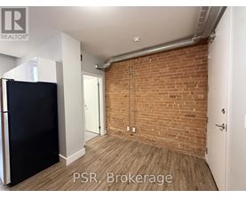 A room with a brick wall and wooden flooring, with a black refrigerator on the left side.