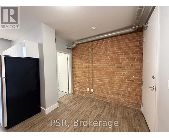 A room with a brick wall and wooden flooring, with a black refrigerator on the left side.