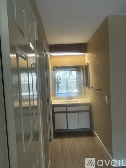 A hallway with a door on the left and a kitchen area with a sink and cabinets on the right.
