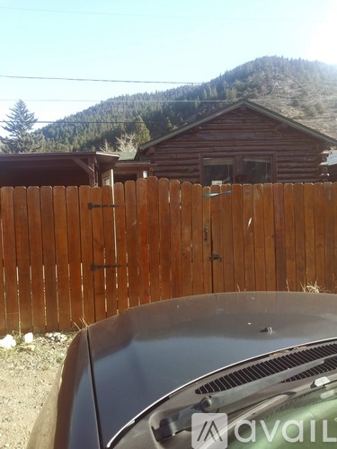 A car is parked in front of a wooden fence with a house and trees in the background.