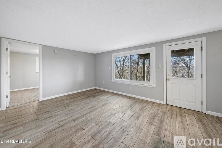 A room with wooden flooring and two windows, one of which has a view of trees.