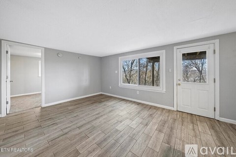 A room with wooden flooring and two windows, one of which has a view of trees.