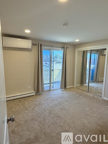 A room with a carpeted floor, a wall-mounted air conditioner, and sliding glass doors leading to a balcony.
