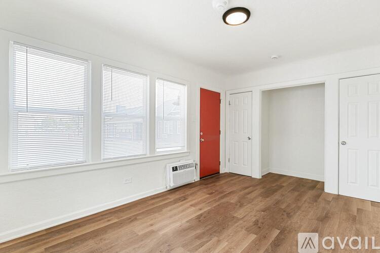 A room with wooden floors and white walls, with a window covered by blinds, a red door, and a white door.