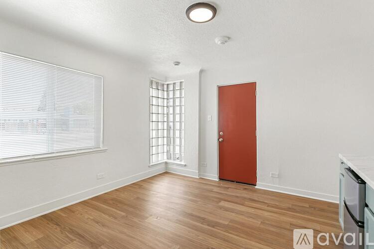 A room with a wooden floor, a red door, and a window with blinds.