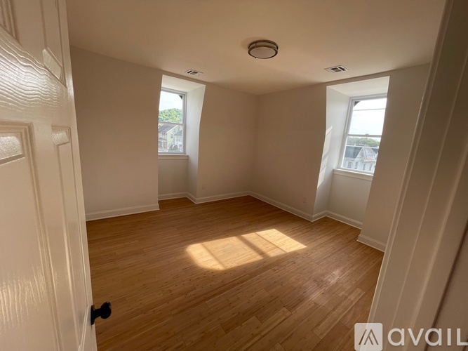 A room with wooden flooring and white walls, with a door on the left and a window on the right.