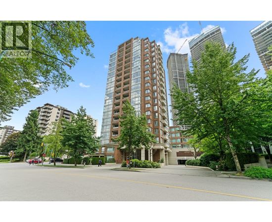 A tall building with a brown facade stands in the middle of a street with trees and other buildings in the background.