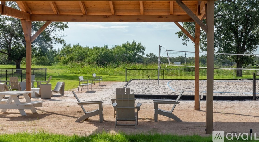 A wooden pavilion with a picnic table and chairs is surrounded by a fence and trees.