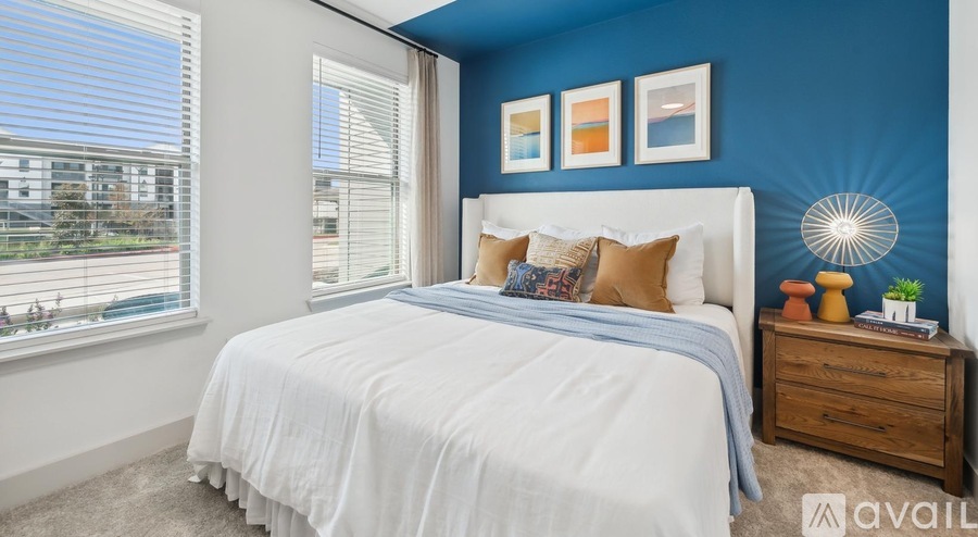A bedroom with a white bed, a window with blinds, and a wooden nightstand with a lamp and a plant on it.