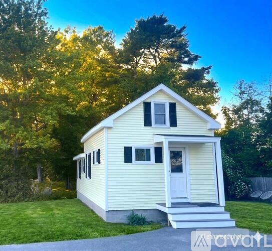 A small yellow house with a white door and windows is surrounded by greenery.