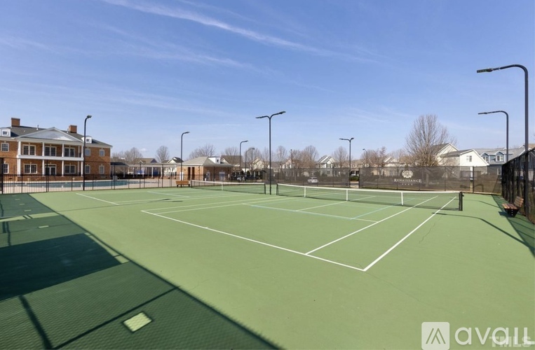 A tennis court with a green surface and white lines, surrounded by a fence and buildings in the background.