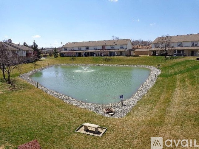 A pond with a fountain in the middle of a grassy area with houses in the background.