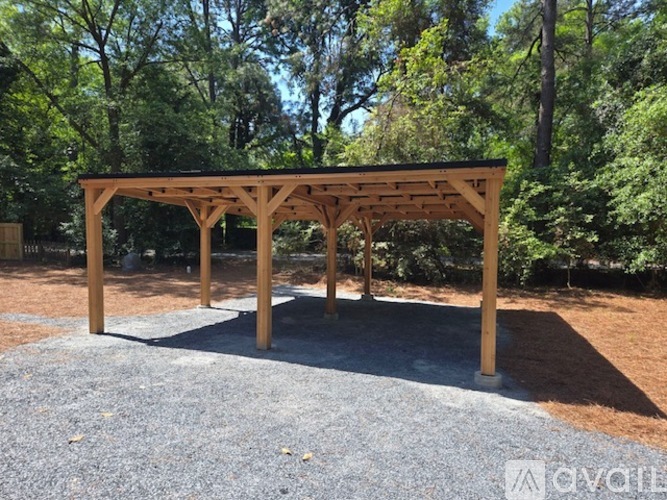 A wooden pergola is situated in a gravel area with trees in the background.