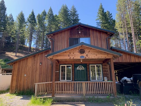 A wooden cabin with a green door and windows is surrounded by trees.
