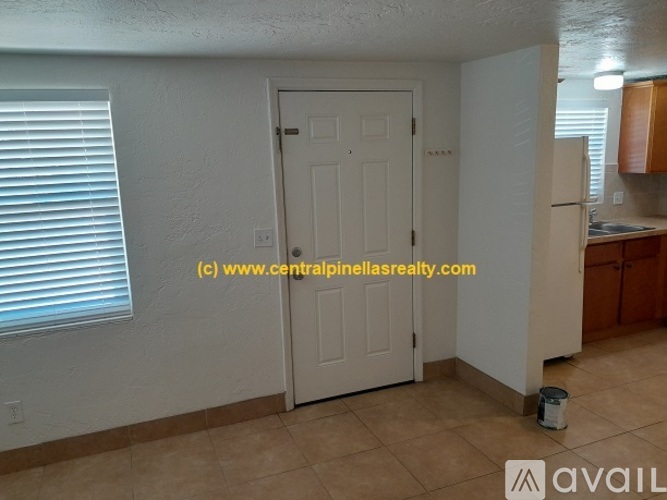 A room with a white door, a window with blinds, and a kitchenette with wooden cabinets.