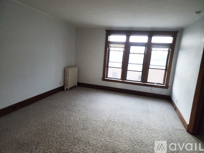 A room with a carpeted floor, a window with a view of a building outside, and a radiator on the wall.