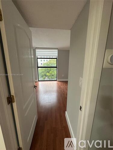 A hallway with a wooden floor and white walls leading to a door with a view of trees outside.