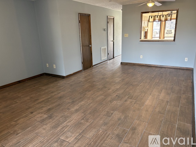 A room with wood flooring and a window with a sign that says "FOR RENT".