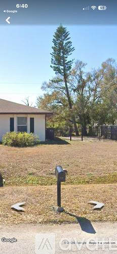 A mailbox stands in the middle of a grassy area with a house and trees in the background.