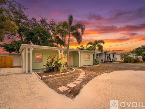 A house with a green exterior is surrounded by a gravel driveway and palm trees.