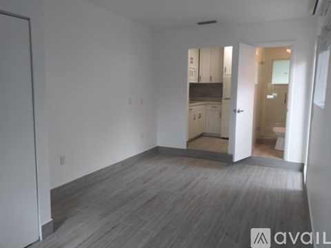 A room with wooden flooring and white walls, leading to a kitchen area.