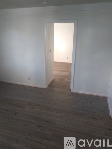A room with wooden flooring and white walls, leading to another room.