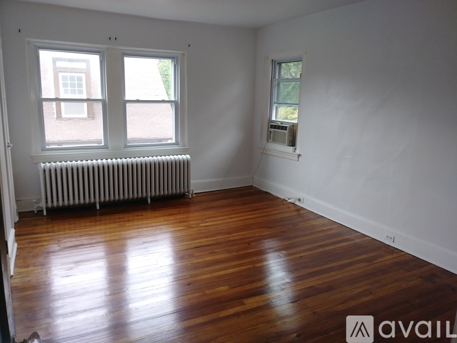 A room with wooden floors and white walls, featuring a window with a view of a building and a radiator.