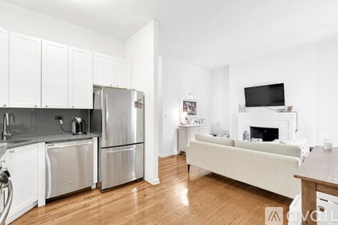 A kitchen with white cabinets and a refrigerator, a white sofa, and a fireplace in the living room.