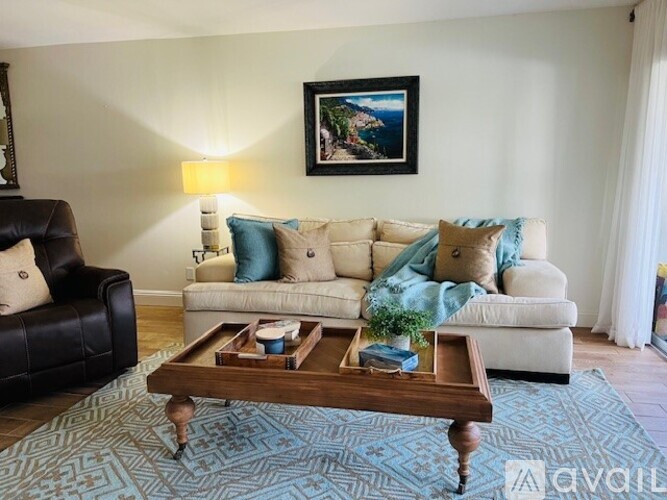 A living room with a black leather chair, a white sofa with blue and beige cushions, a wooden coffee table, and a painting on the wall.
