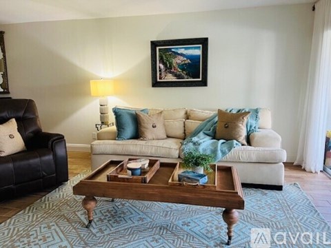 A living room with a black leather chair, a white sofa with blue and beige cushions, a wooden coffee table, and a painting on the wall.