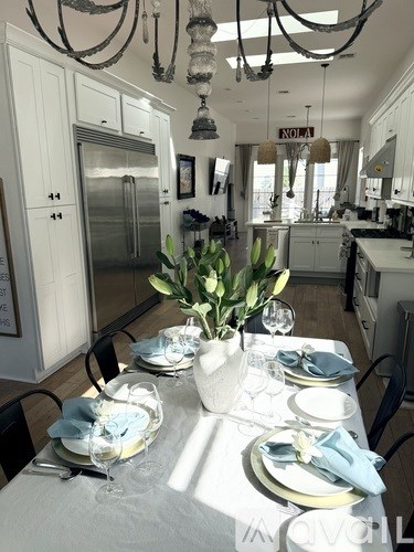 A dining table set with plates and glasses is in the foreground of a well-lit kitchen.