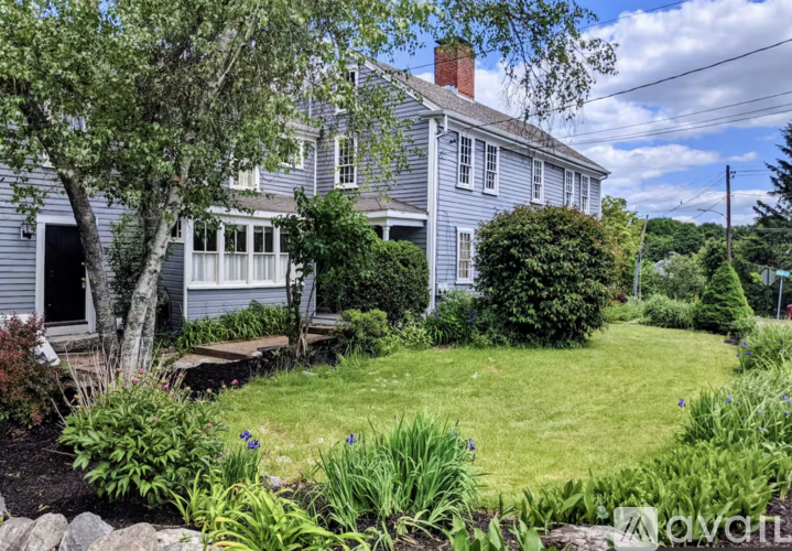 A house with a grey exterior and a white window is surrounded by a garden with a variety of plants and flowers.