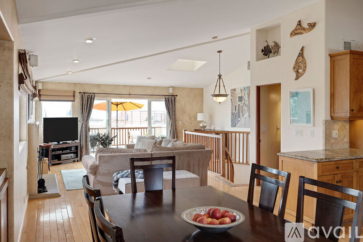 A dining room with a wooden table and chairs, a bowl of fruit on the table, and a TV in the background.
