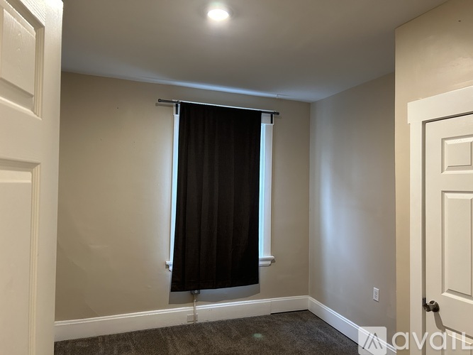 A room with a carpeted floor, a window with a black curtain, and a white door.