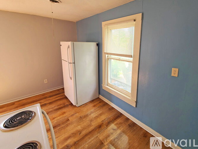 A room with a white fridge, a window with blinds, and a wood floor.