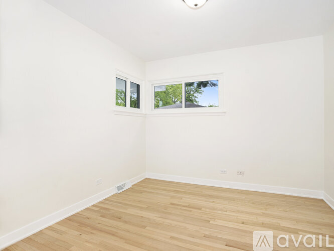 A room with wooden flooring and two windows showing trees and sky.