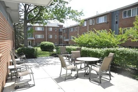 A patio with chairs and a table is surrounded by a brick wall and a green hedge.