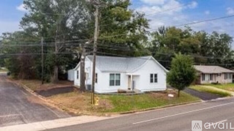 A white house with a grey roof is situated on a street with a tree in front of it.