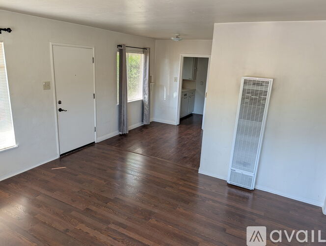 A room with wooden floors and white walls, with a door on the left and a window with blinds on the right.