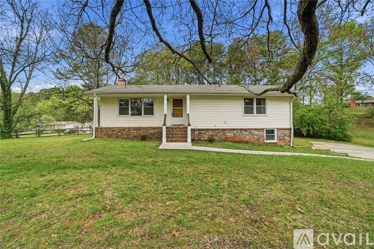 A house with a white front porch and a brown door is surrounded by a grassy lawn and trees.