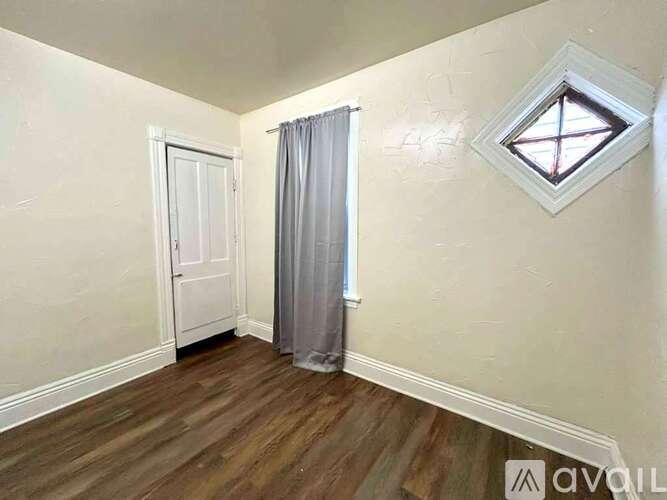 A room with a wooden floor, a white door, a grey curtain, and a window with a diamond-shaped frame.