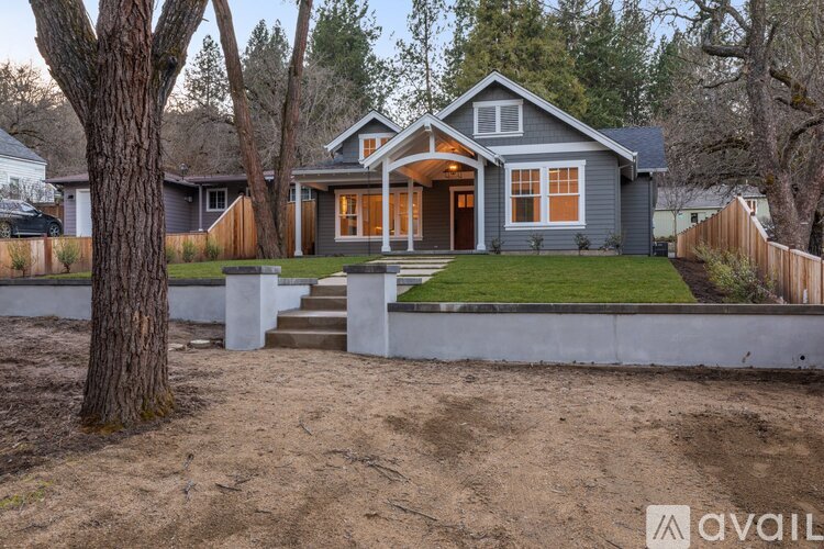A house with a grey exterior and a brown roof is surrounded by a white fence and a tree.