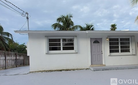 A small white house with a grey door and windows is surrounded by a fence and palm trees.
