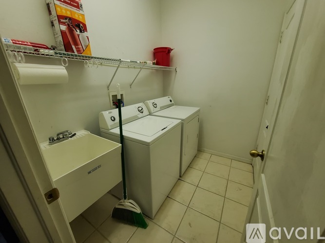 A small white bathroom with a toilet, a trash can, a broom and a shelf with toilet paper.
